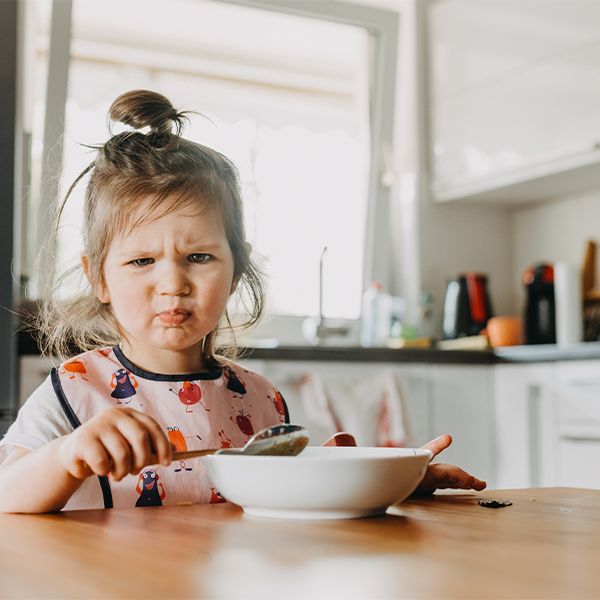 Toddler frowning at a bowl of food at a kitchen table.