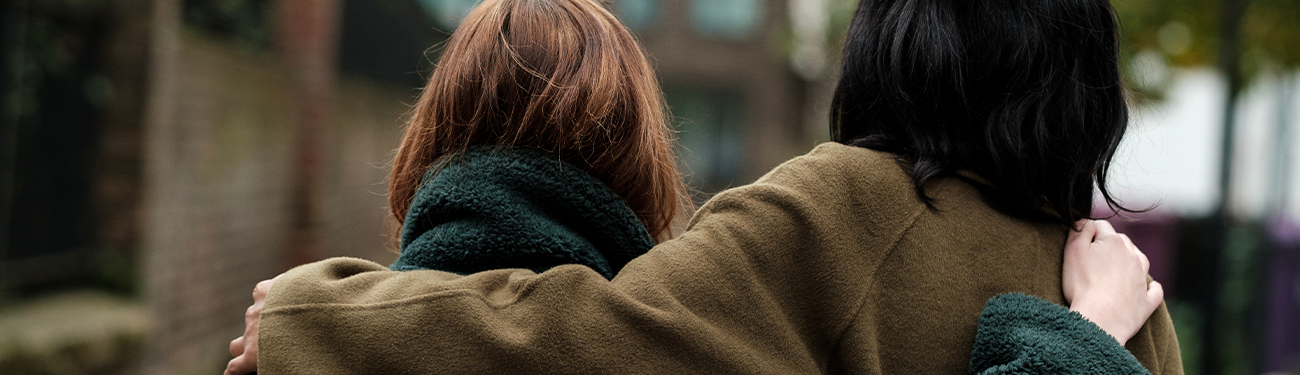 Two women in brown coats walk closely together outdoors, seen from behind.