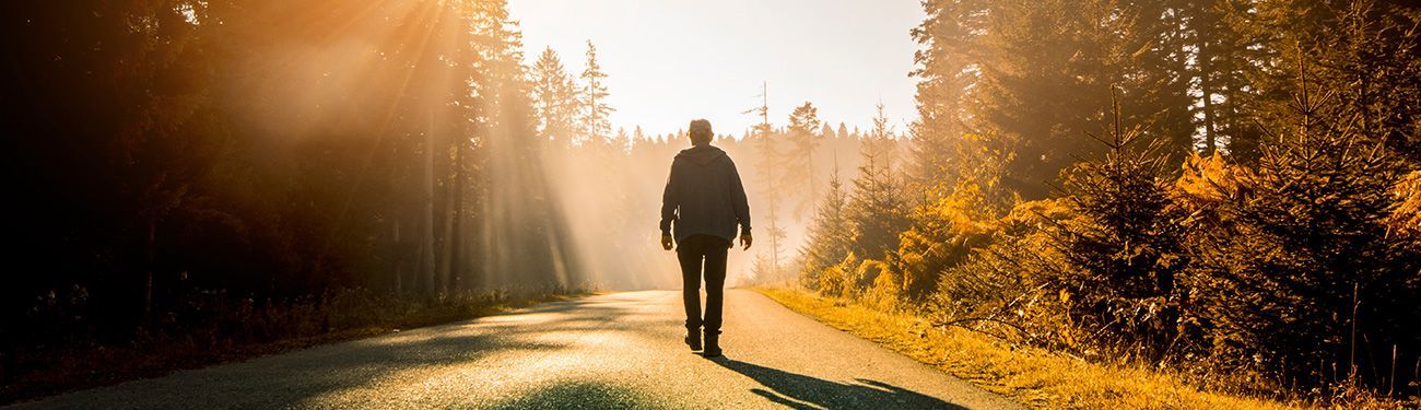 A man is walking down a road in the woods at sunset.