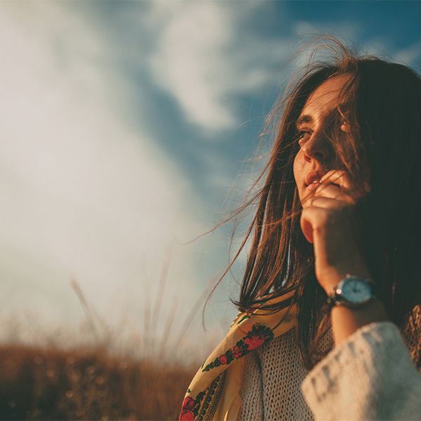 Woman with long hair gazes upward in a windy field under a cloudy sky, hand near her face.