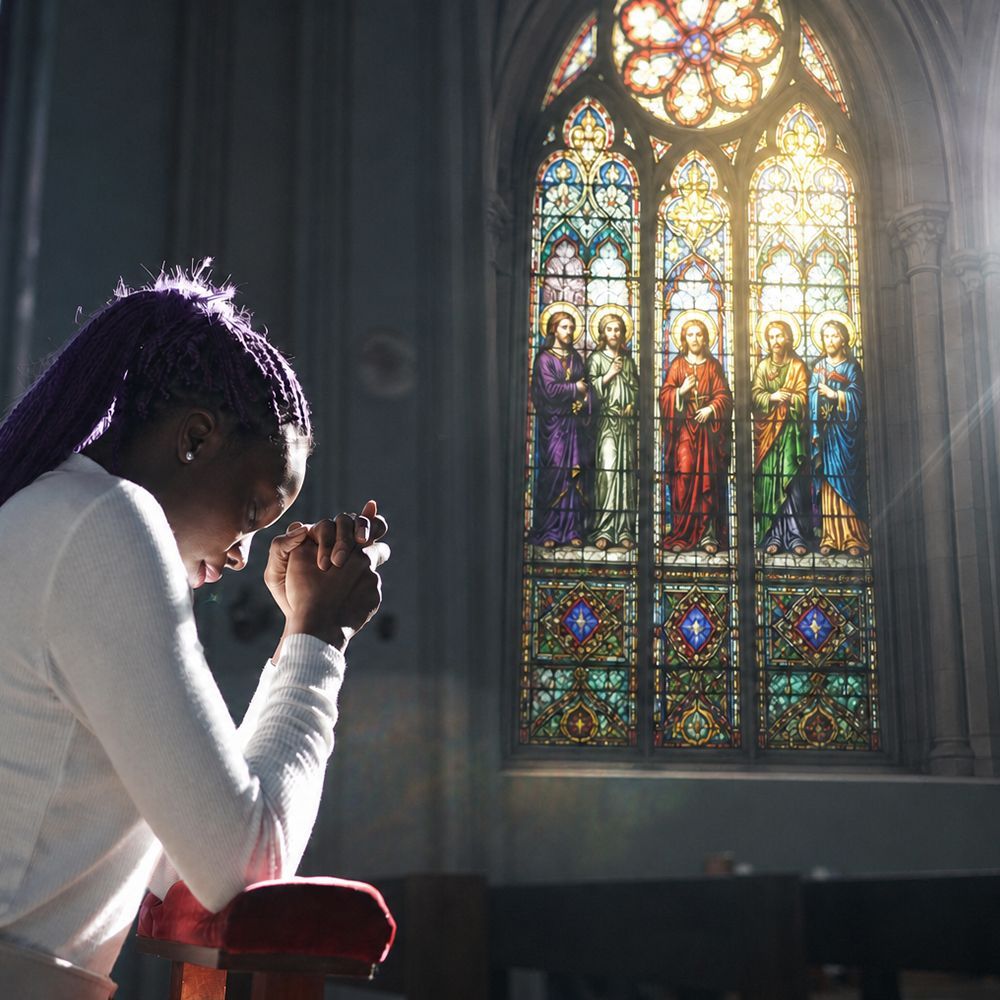 Woman praying beside a stained-glass church window featuring Catholic saints, lit by soft sunlight.