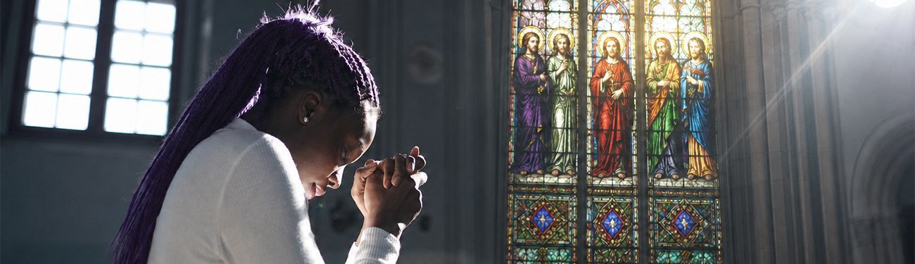 Woman praying beside a stained-glass church window featuring Catholic saints, lit by soft sunlight.