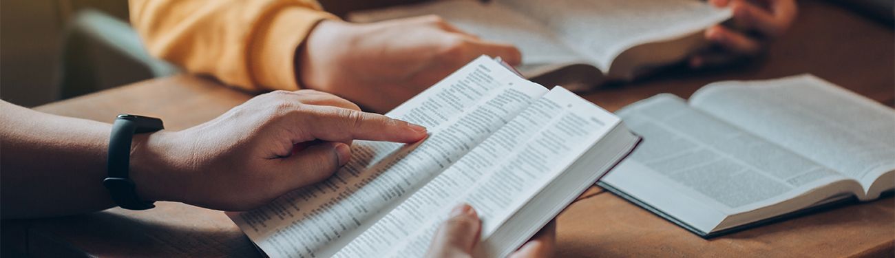 People reading open Bibles at a table, with a close-up of hands turning pages.