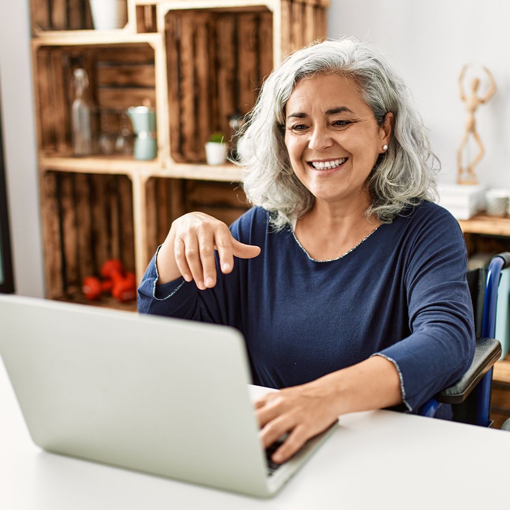 Smiling woman using a laptop at a table in a bright room with wooden shelves behind her