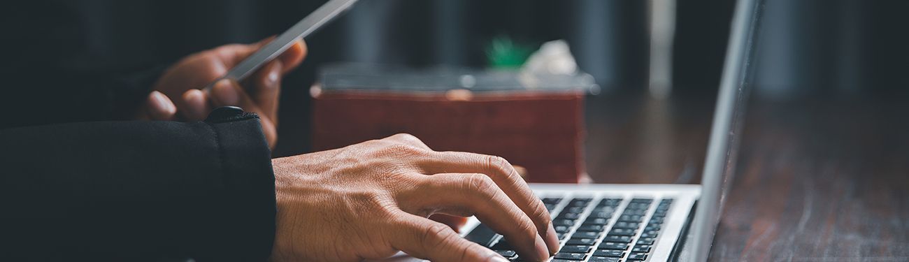 A person is typing on a laptop computer while holding a cell phone.