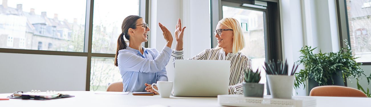 Two women are sitting at a table in front of a laptop computer giving each other a high five.