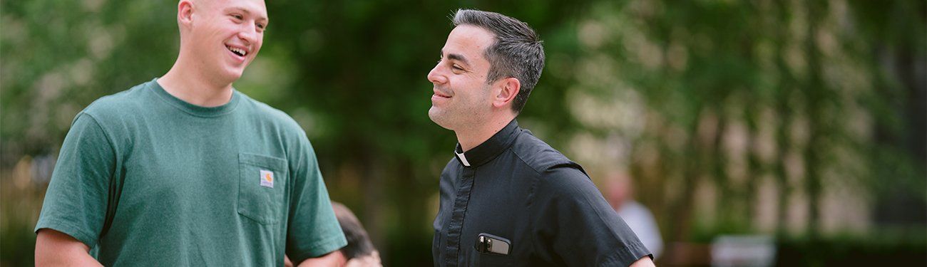 A Preist and a man are standing next to each other in a park talking to each other.