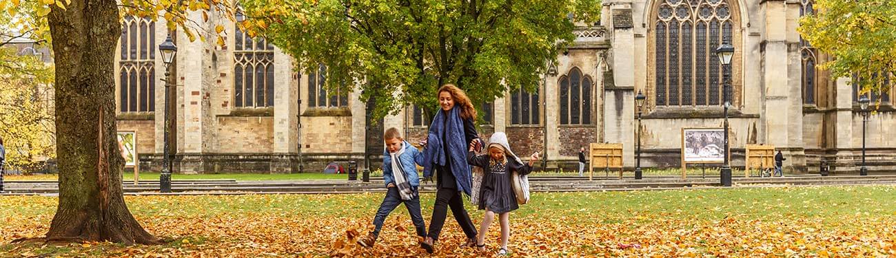 A woman and two children are walking through a park with leaves on the ground.