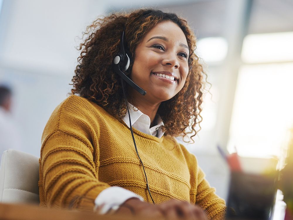 Smiling headset-wearing customer service agent in a mustard sweater at a desk