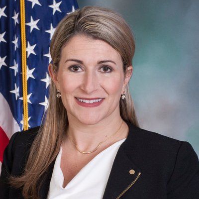 A woman in a black jacket and white shirt is smiling in front of an american flag.