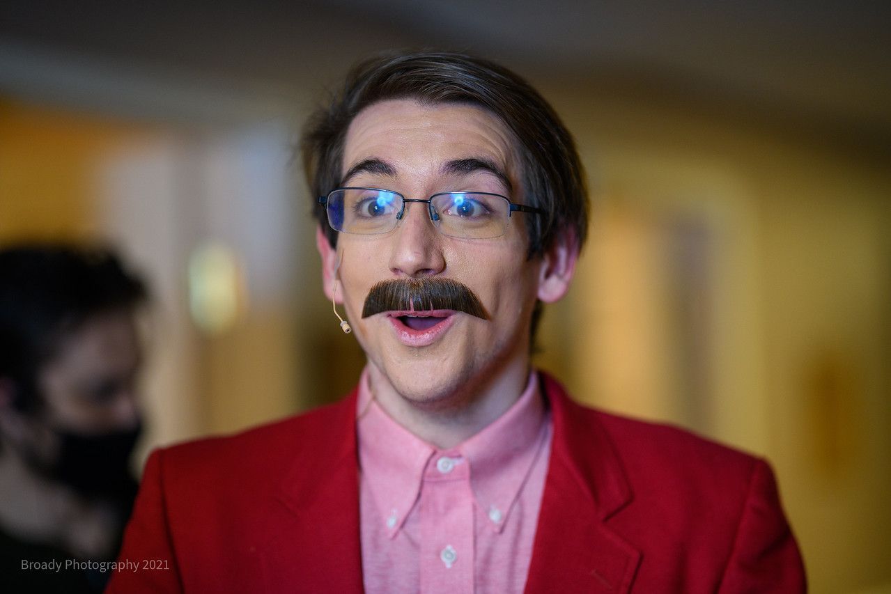 A man with a mustache and glasses is wearing a red jacket and a pink shirt.