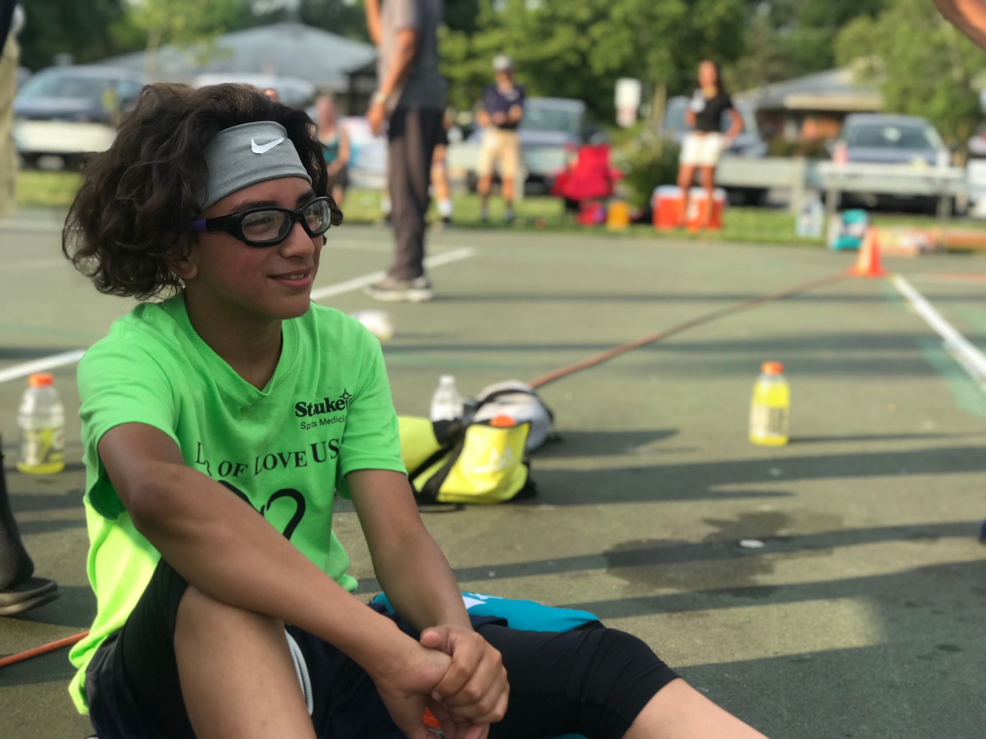 A young boy wearing glasses and a headband sits on a tennis court