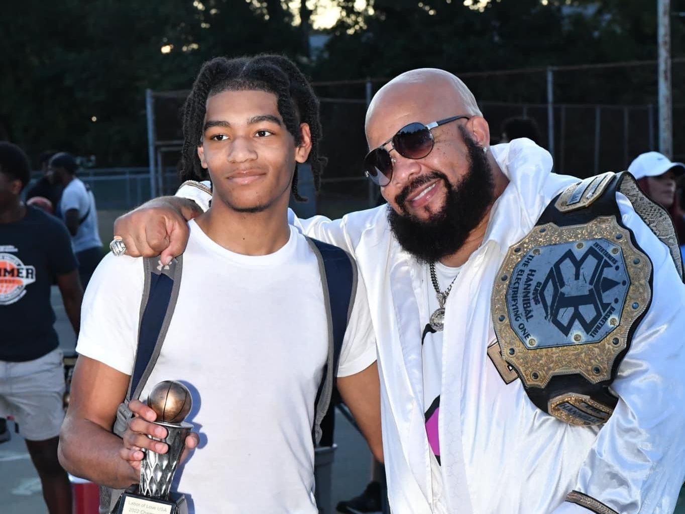 A bald man with a beard is standing next to a young man holding a trophy.