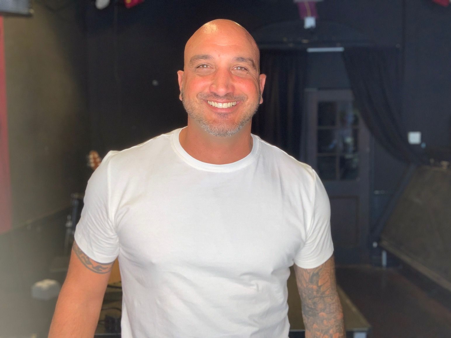 A bald man is wearing a white t-shirt and smiling.
