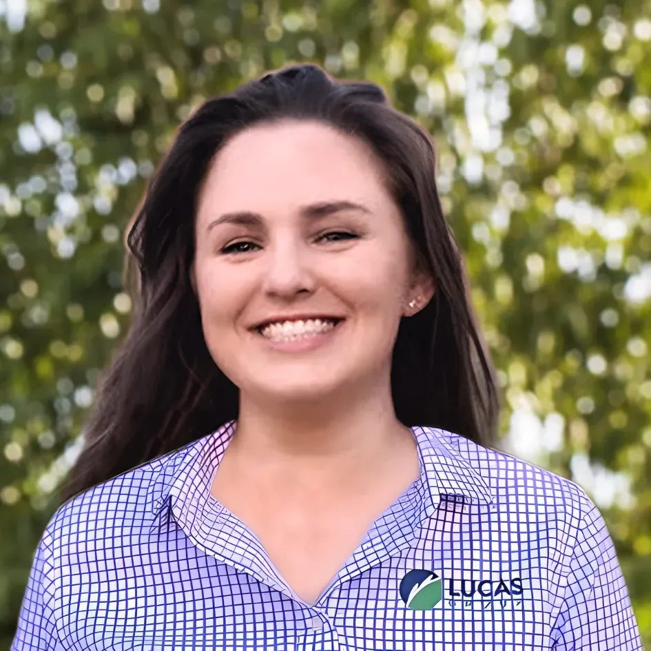 Woman smiling, wearing a blue patterned shirt with Lucas logo. Outdoor setting with green foliage.