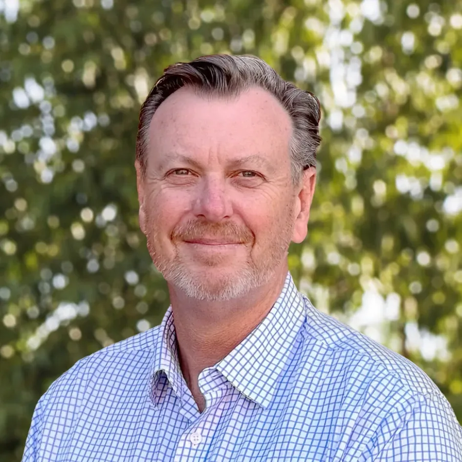 Man with graying hair smiles, wearing a blue and white checkered shirt outdoors.