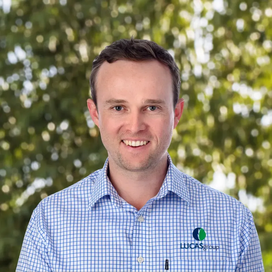 Man smiling, wearing a blue plaid shirt with a logo, set against a blurred green background.