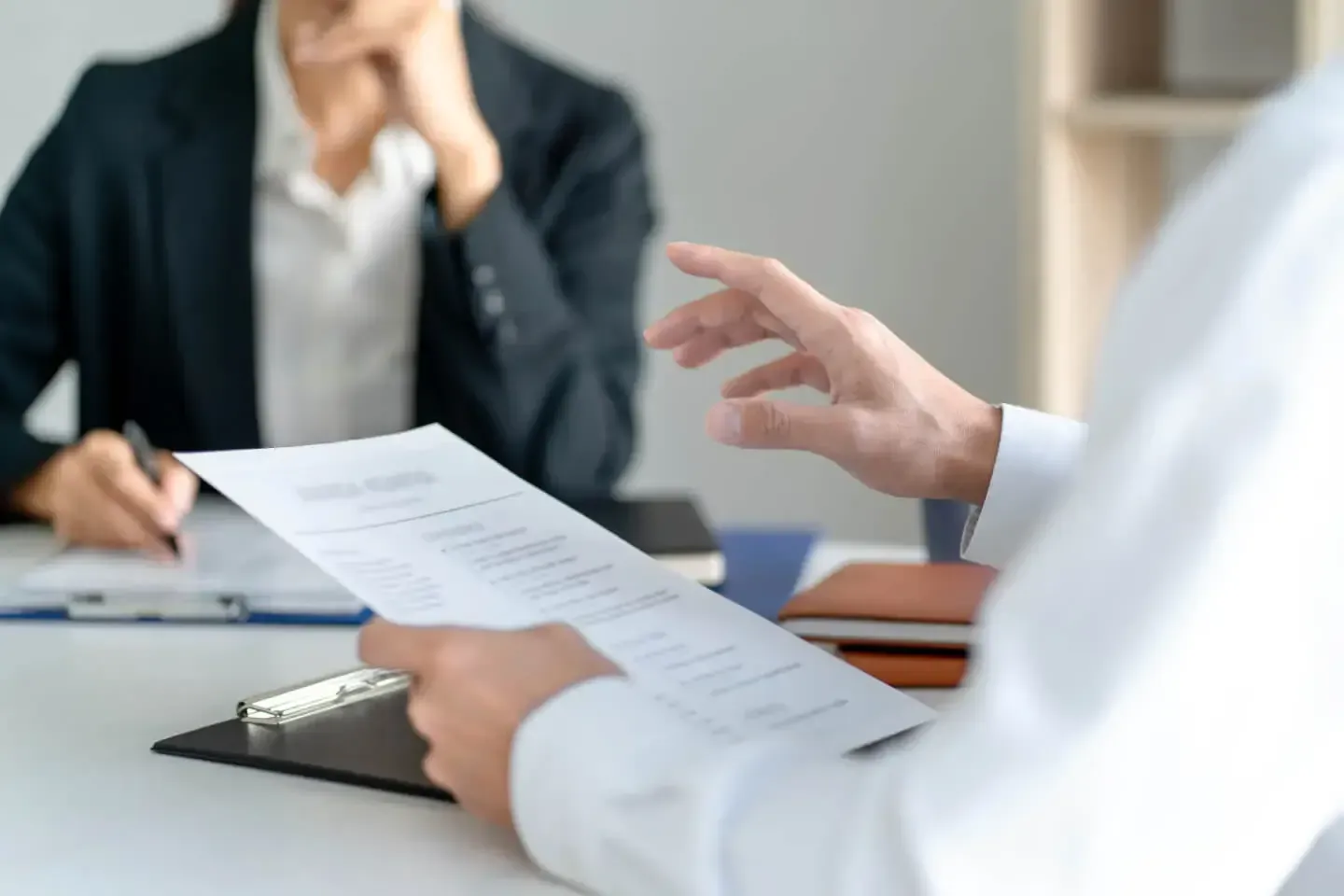 Person reviewing a document during a meeting, gesturing with hand; another person in background takes notes.