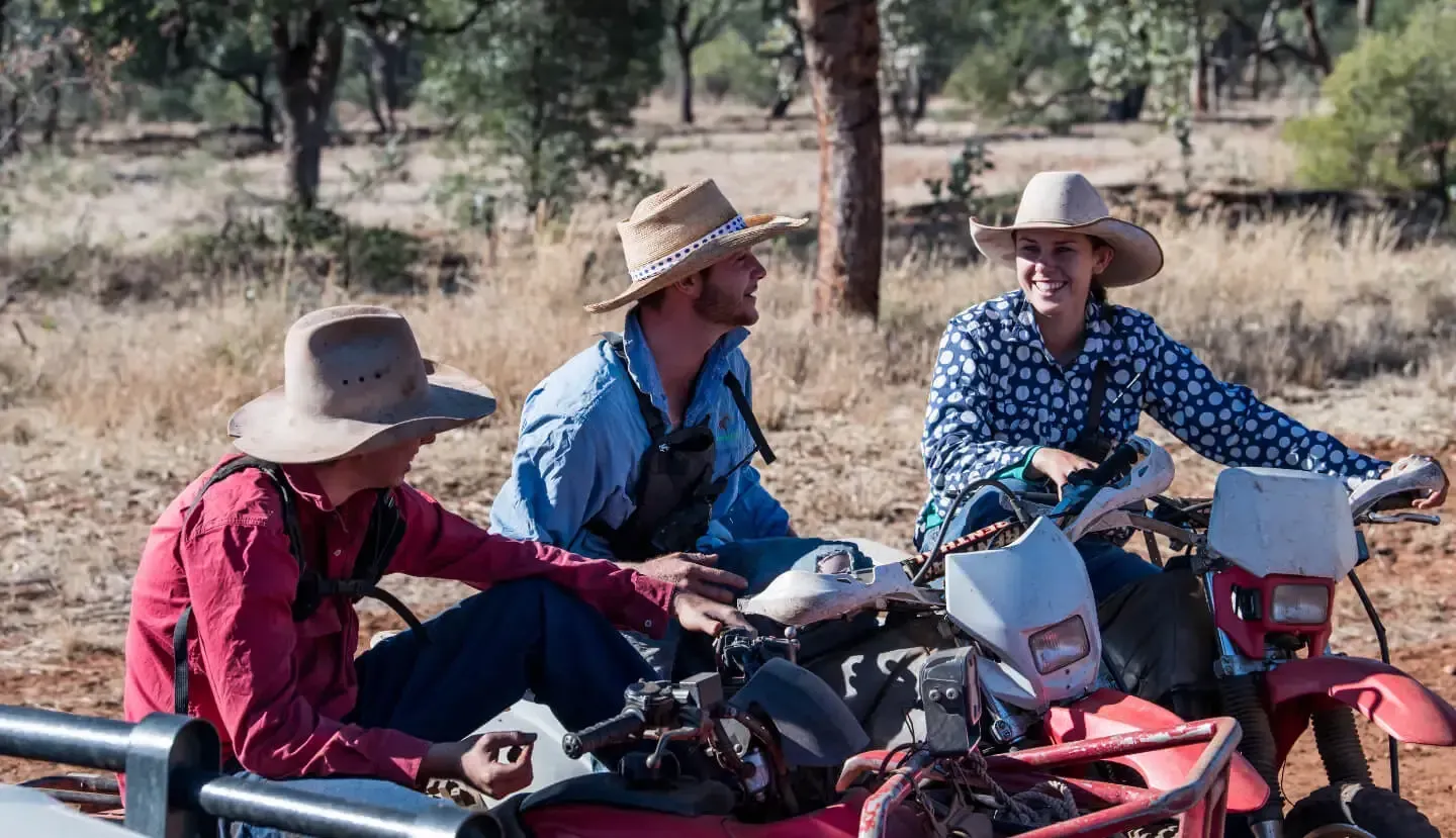 Three people wearing hats sit on a red motorbike in a dry, outdoor environment, smiling.