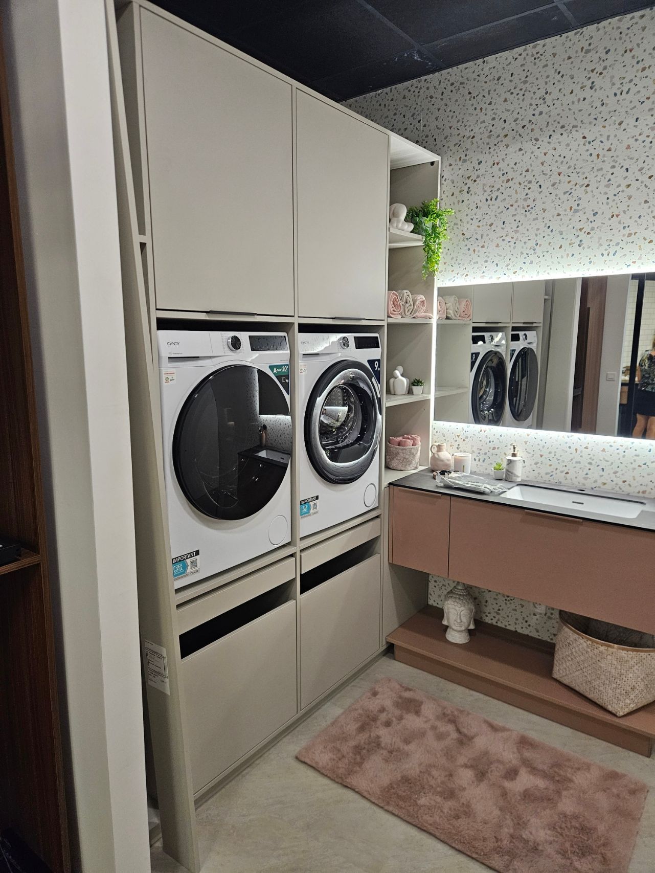 Petite buanderie avec lave-linge et sèche-linge superposés, miroir, évier et tapis rose.