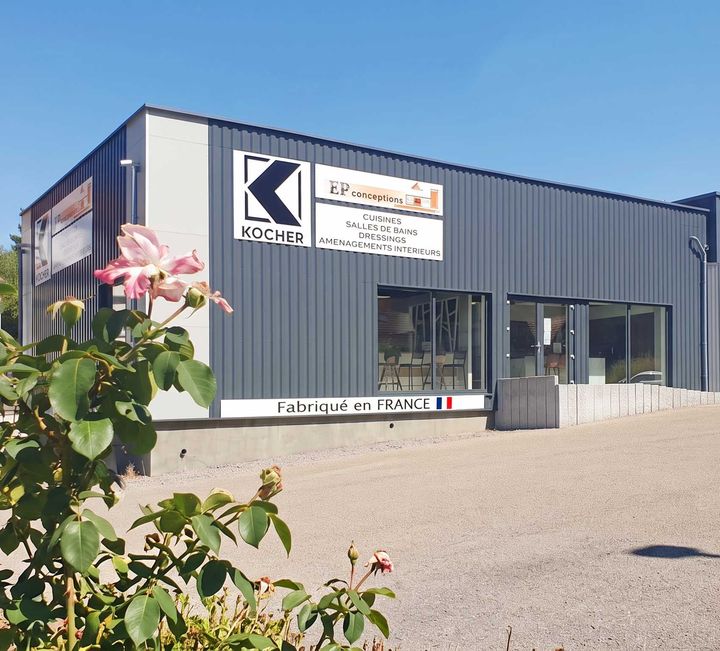 Bâtiment industriel avec enseigne Kogher, bardage gris foncé et fleurs au premier plan sous un ciel bleu clair.