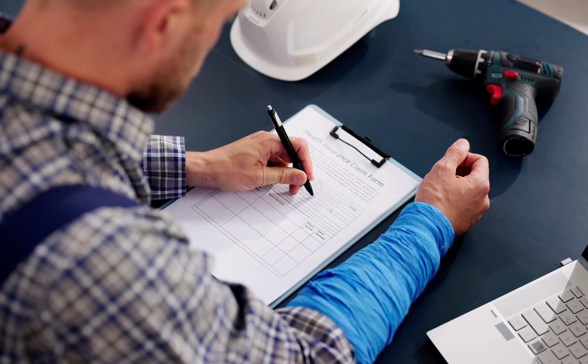 Construction worker writing on a clipboard; desk with hard hat, drill, and laptop.