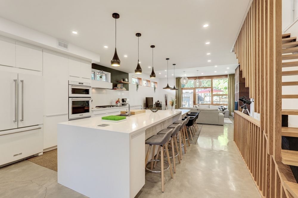 Modern white kitchen with a large island and bar stools, leading to a living area and staircase.