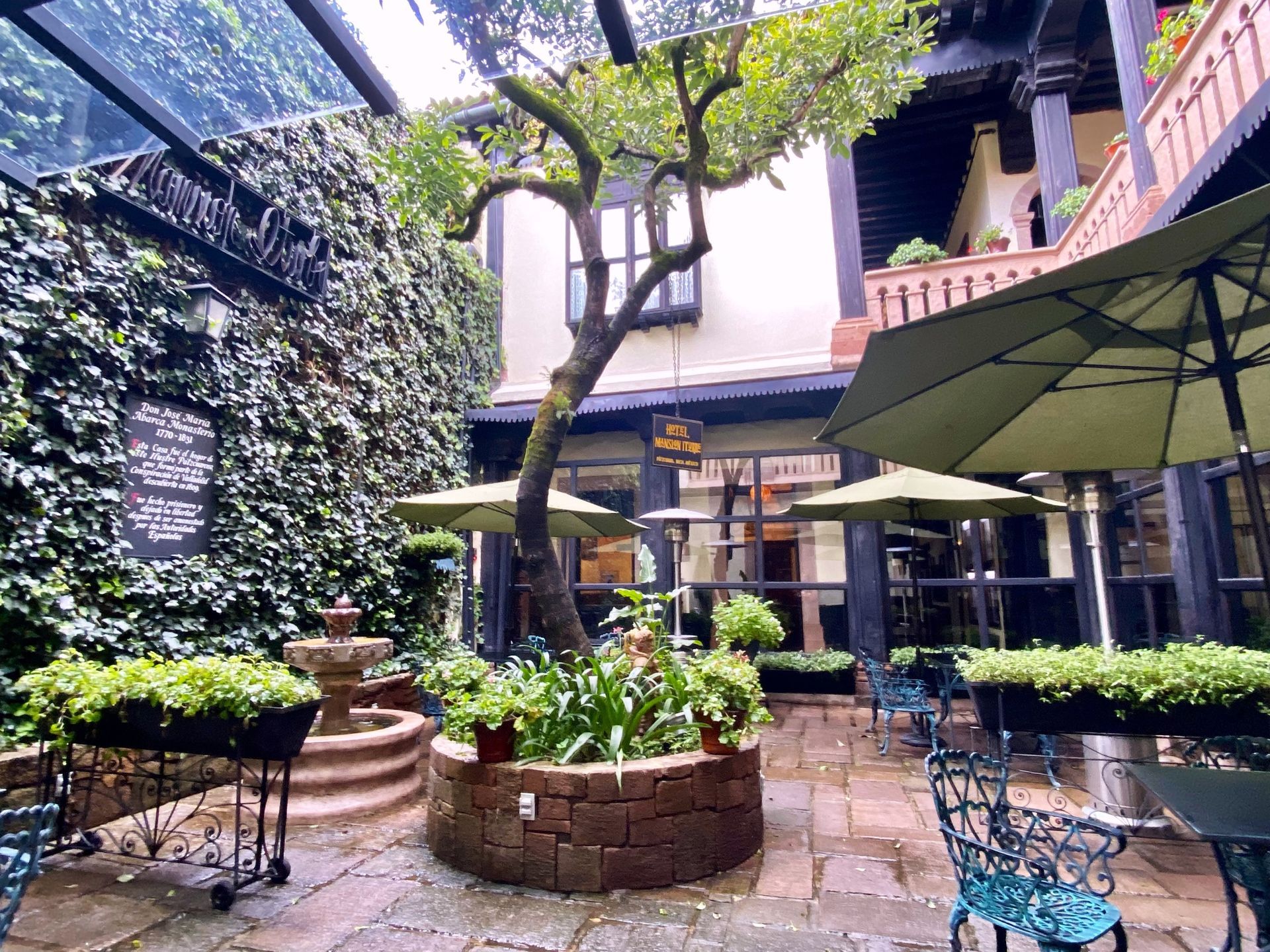 A courtyard with a brick floor, tables, umbrellas, and a fountain, surrounded by a building with ivy and an overhanging tree.
