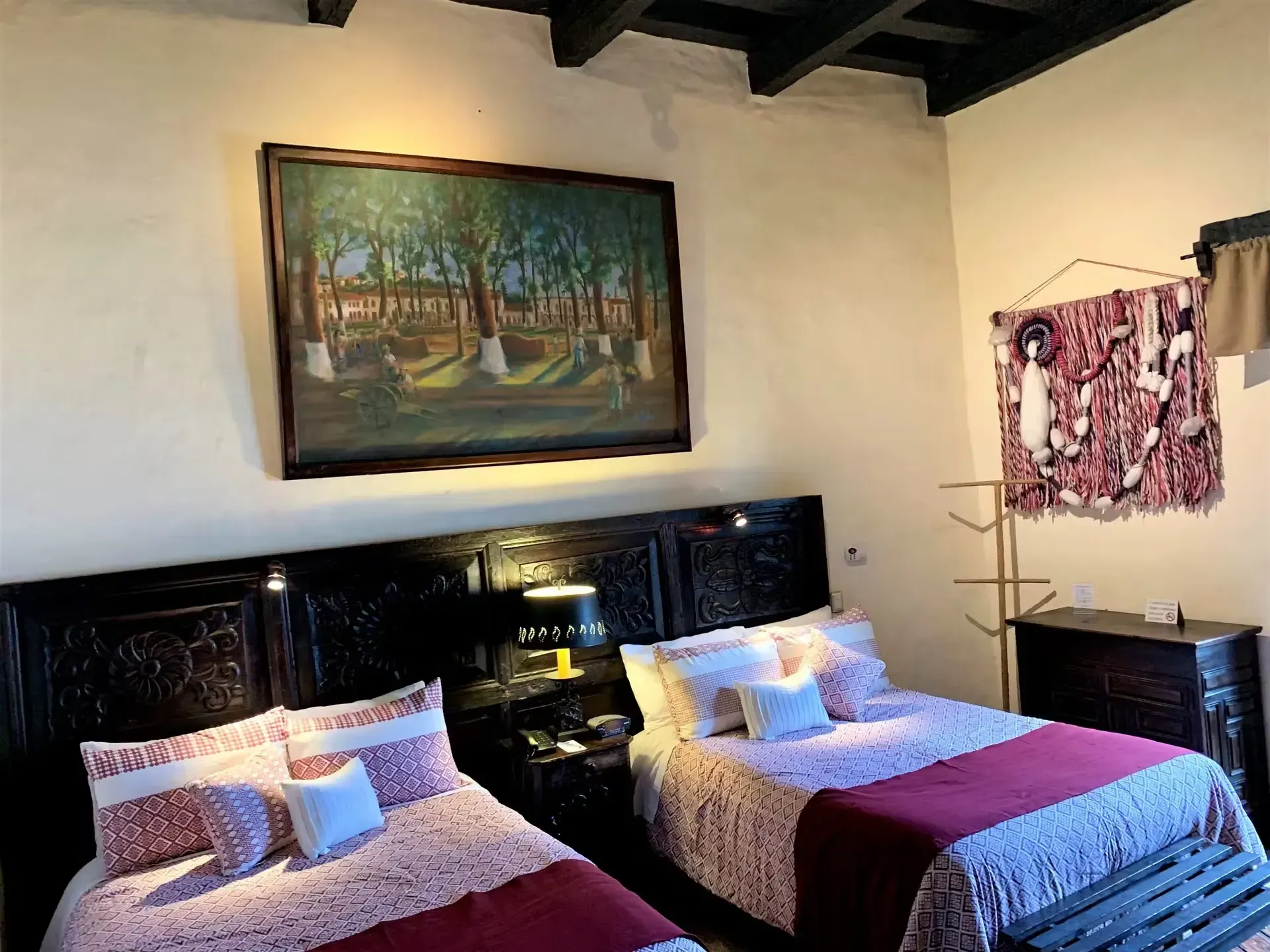 Two beds in a warmly lit room with carved wooden headboards. A painting hangs above, and a textile tapestry to the right.