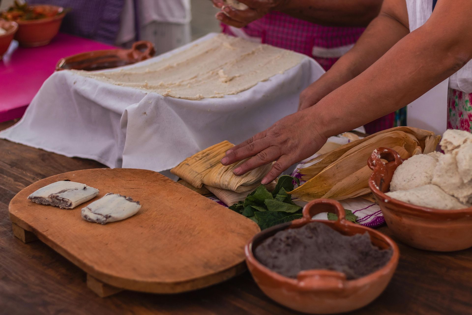 Person preparing Mexican food.  Includes tortillas, tamales, beans, and other ingredients on a wooden board and table.