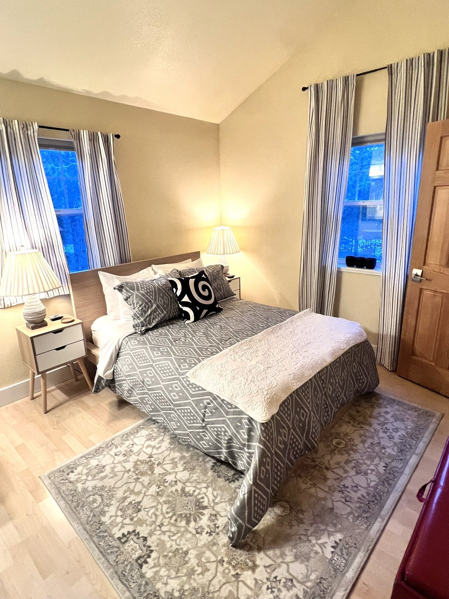 A bedroom with a bed , nightstand , rug and two windows.