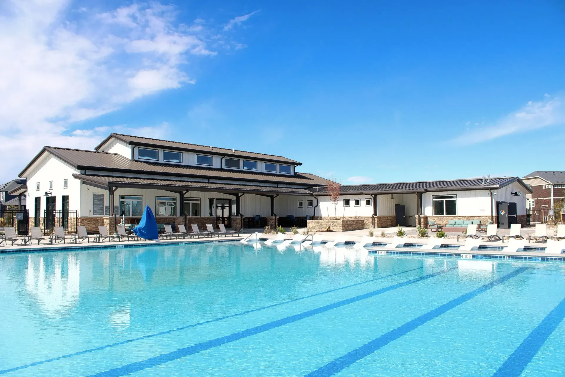 Outdoor swimming pool with clear blue water, lounge chairs, and modern clubhouse against a bright blue sky. 