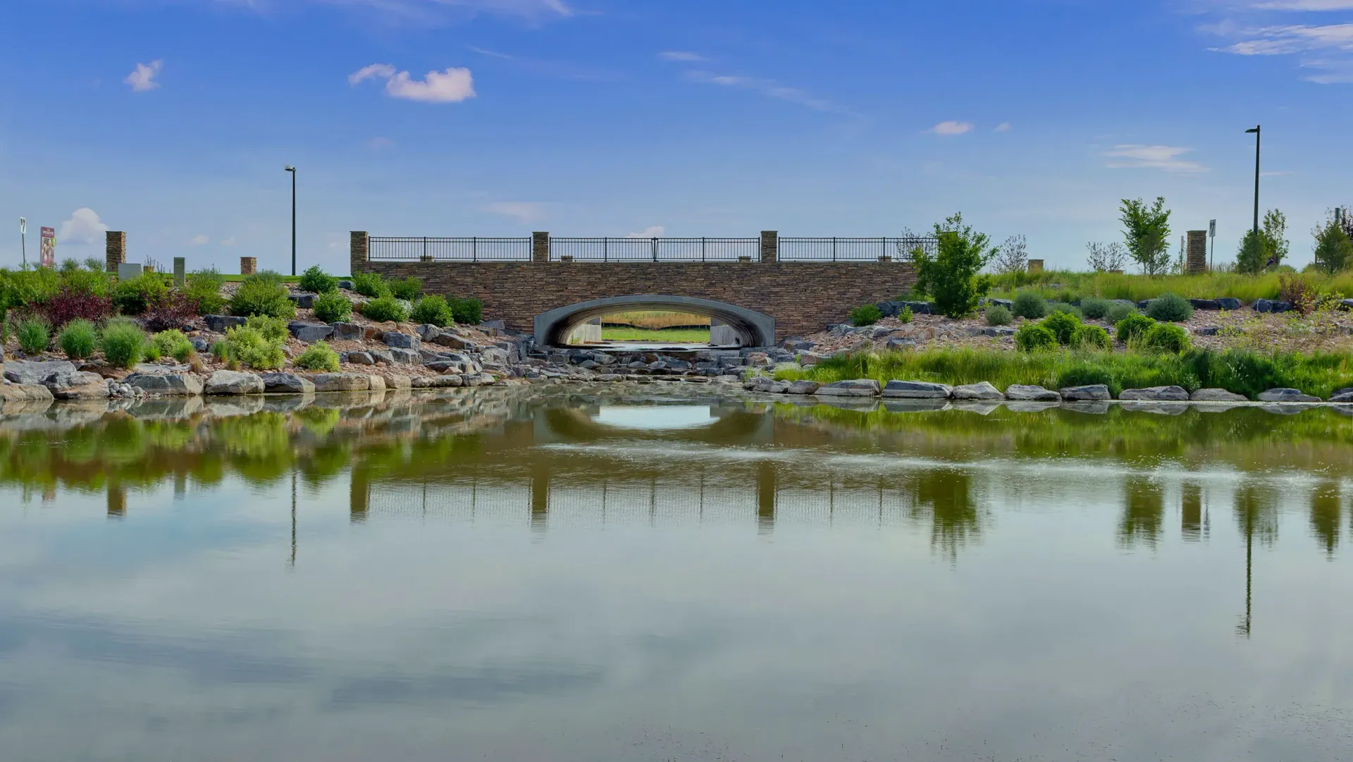 A small stone bridge spans over a calm pond, with grassy banks and scattered bushes.