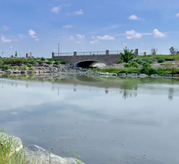 A serene pond reflects a stone bridge and a partly cloudy blue sky.