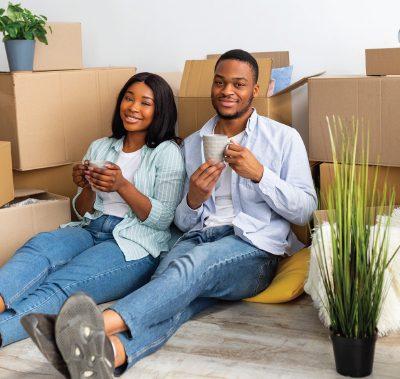 Couple seated on floor, surrounded by moving boxes, drinking from mugs, smiling.