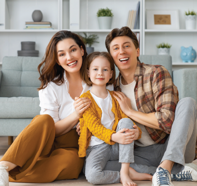 Family of three smiling, posing on a floor in front of a couch.