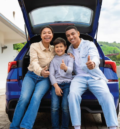 Family of three with thumbs up, seated in blue car trunk. Outdoors, smiling.
