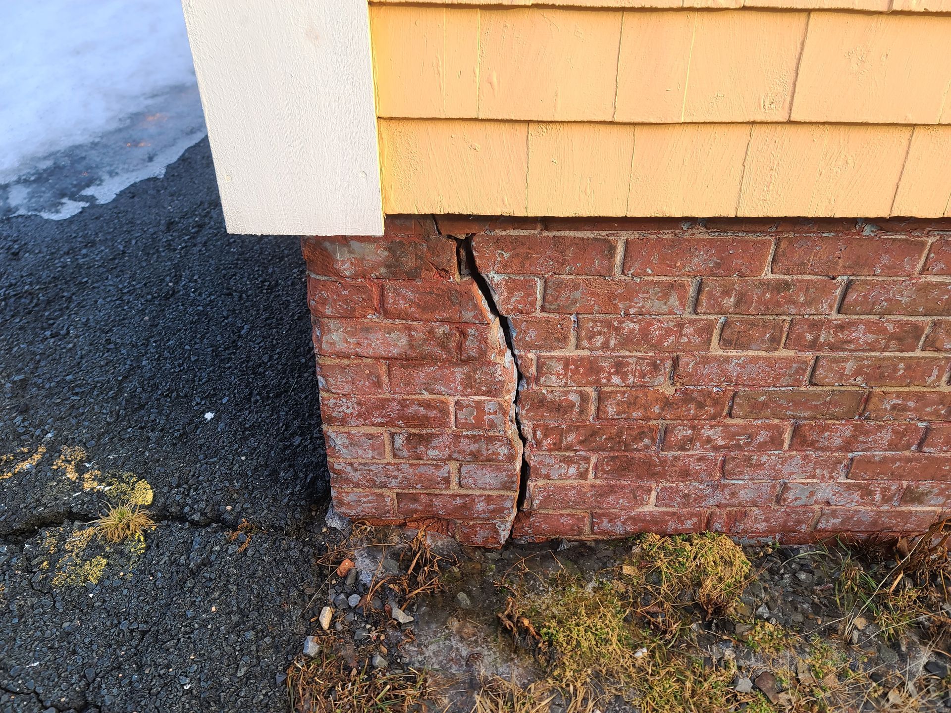 Cracked red brick foundation of a building next to a paved surface. Snow and vegetation visible.