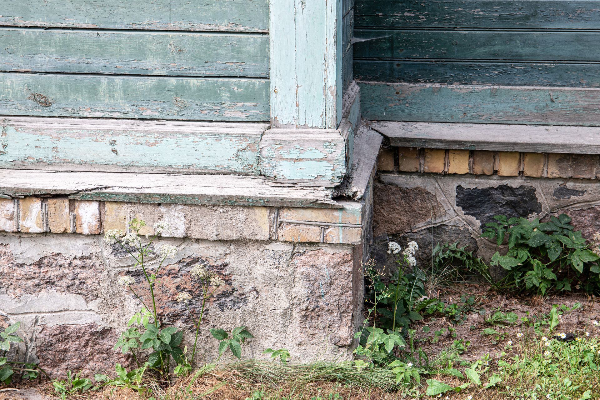 Old building foundation showing deteriorating paint and brickwork, with some weeds.
