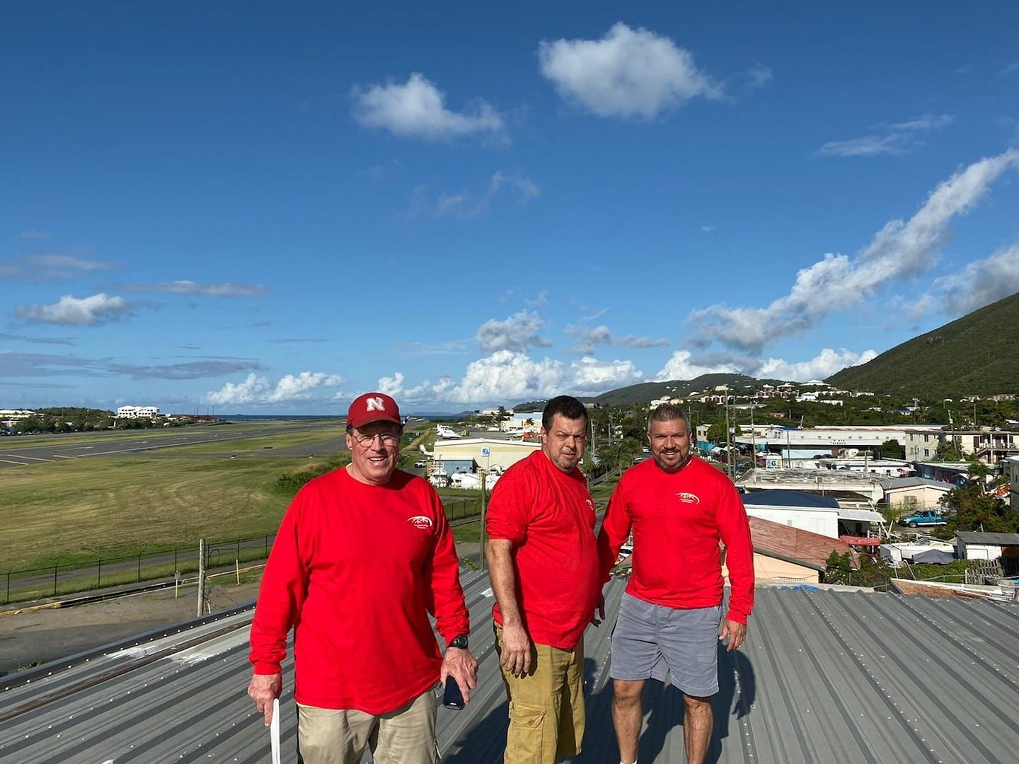 Three people in red shirts standing on a rooftop with a coastal town and hills in the background