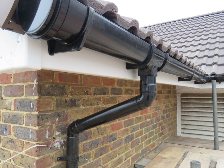 Black gutter downspout on brick house wall beneath tiled roof eaves