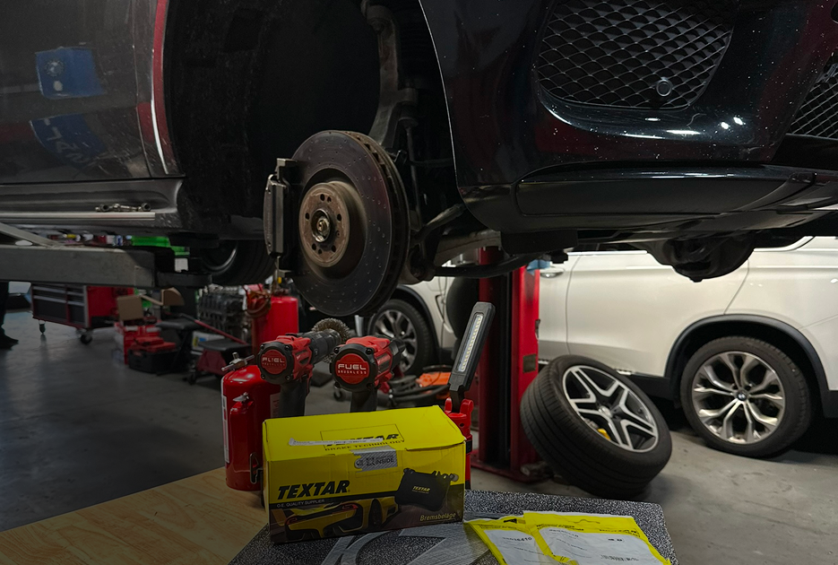 Black SUV lifted on a garage hoist, with removed wheels and parts on a workbench below.