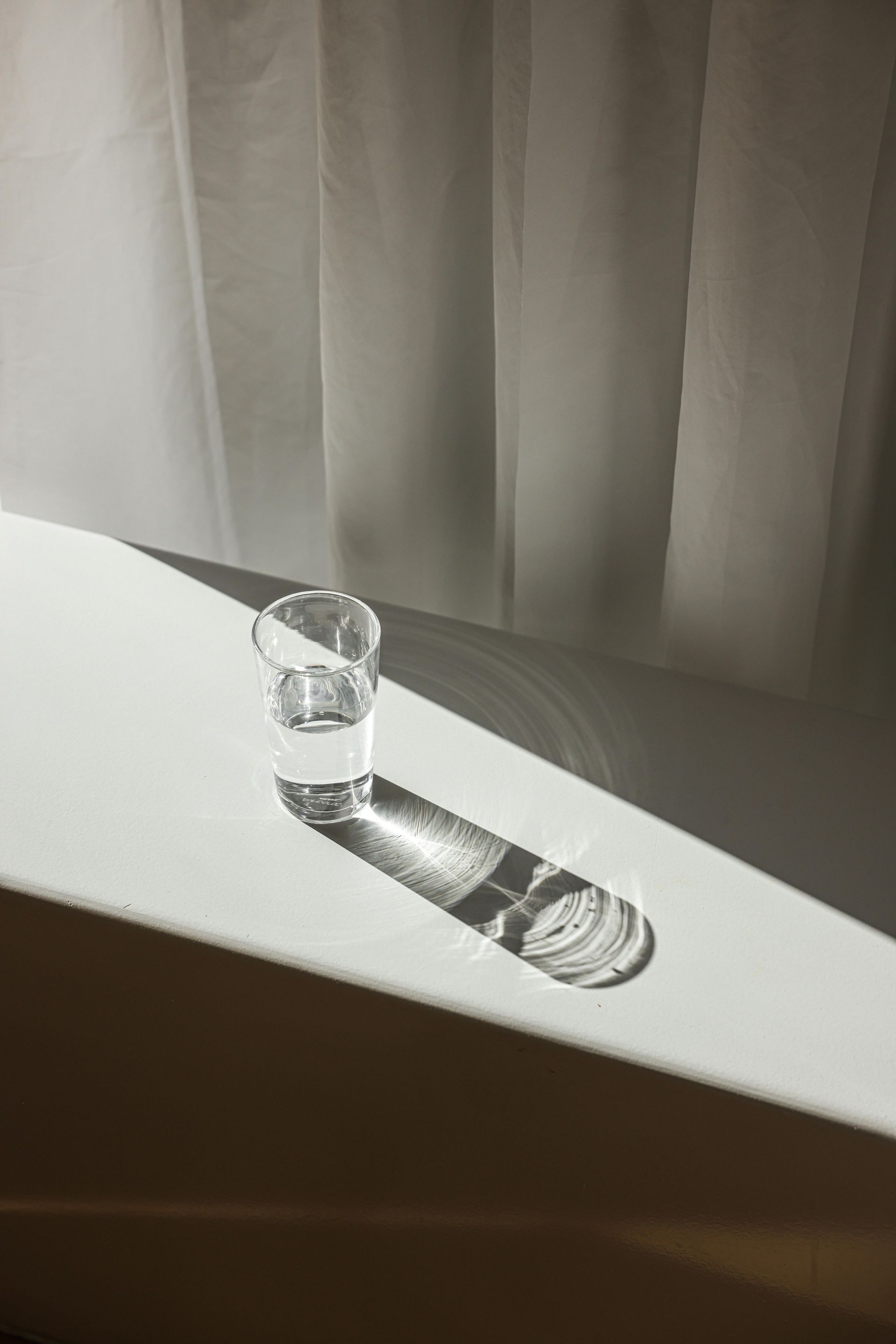 A glass of water is sitting on a white table next to a window.