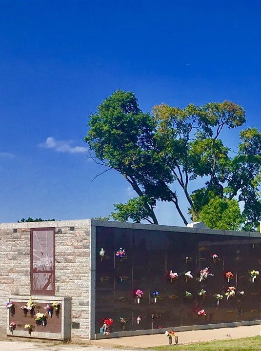 Modern mausoleum with flower tributes beside a tree under a bright blue sky