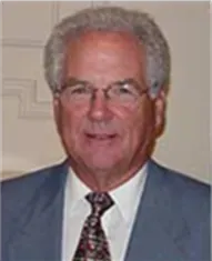 Portrait of a man in a light blue suit and patterned tie, smiling against a beige background
