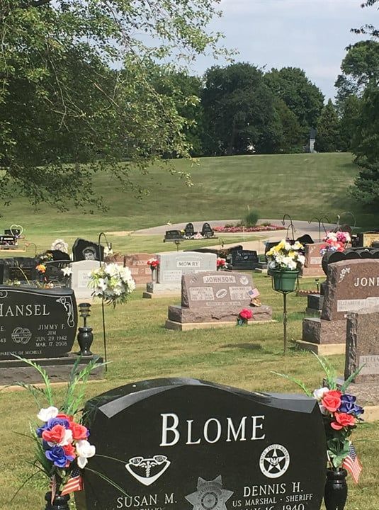 Gravestones in a green cemetery, with a black Blome headstone and trees on a grassy hill.