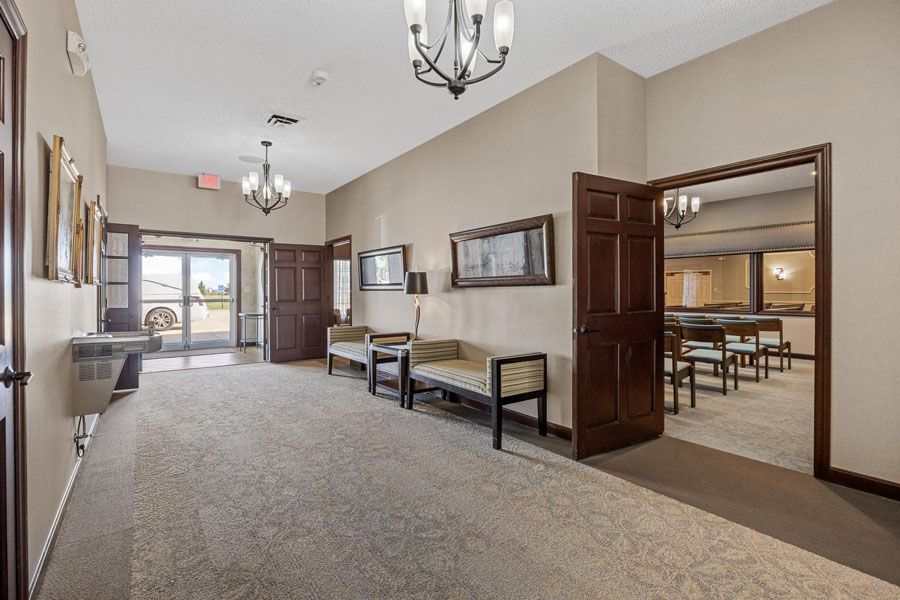 Bright hallway with carpet, benches, chandeliers, and open doors to a dining room and lobby area.
