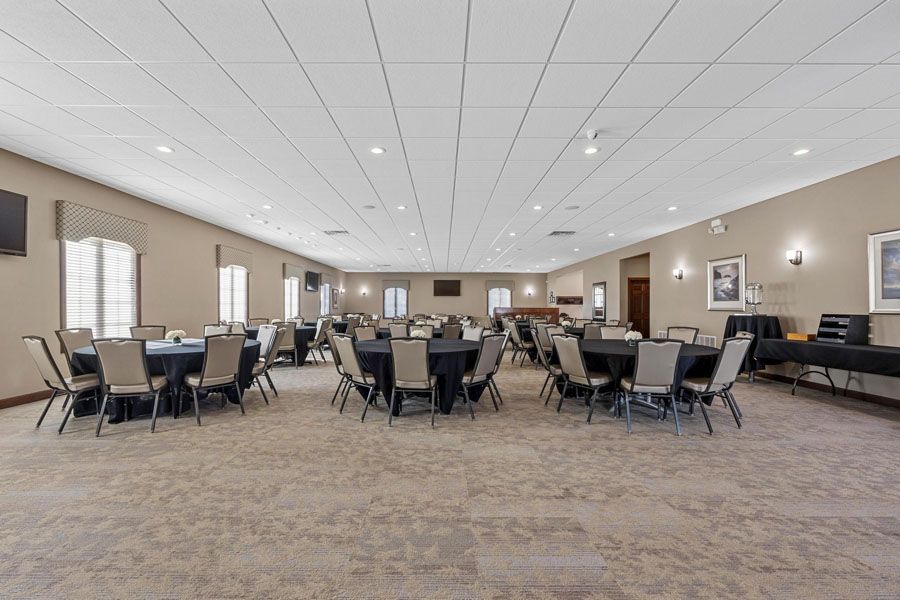 Spacious banquet hall with round tables, black chairs, beige walls, and carpeted floor under recessed lighting