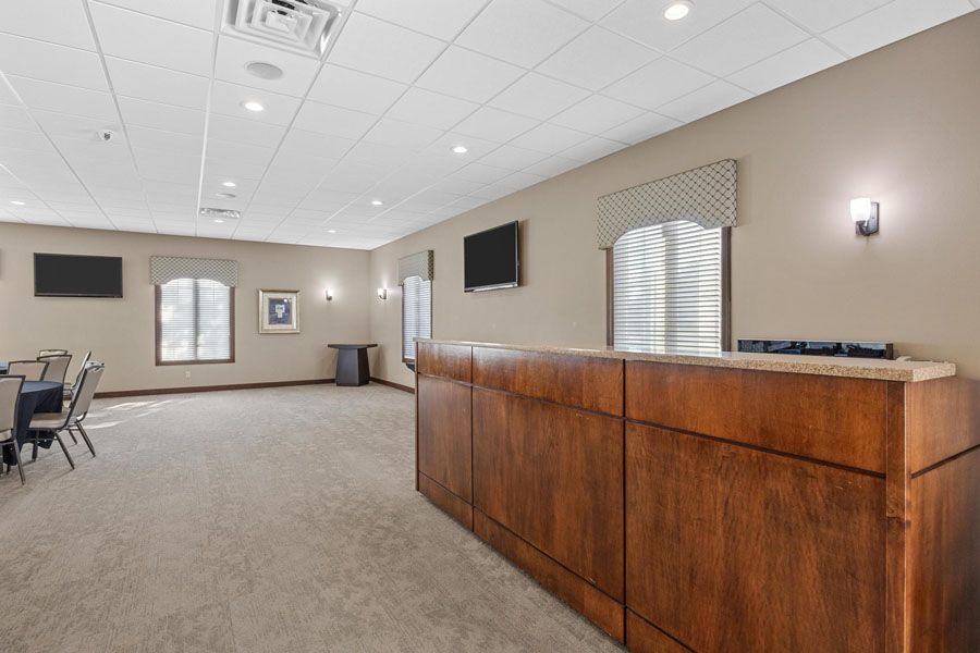 Empty modern lobby with carpeted floor, wooden reception desk, wall-mounted TVs, and tables by windows