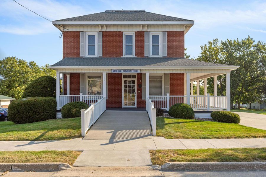 Two-story red brick house with white porch and ramp, front-facing on a sunny suburban street
